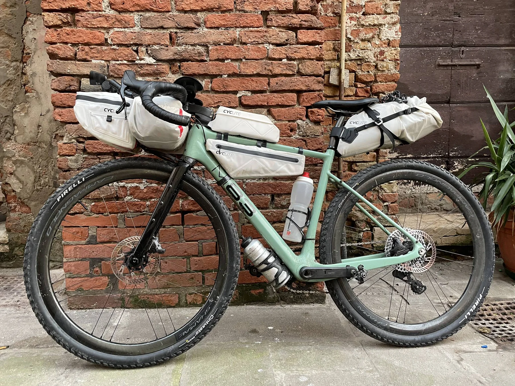 Loaded gravel bike with white bikepacking bags in front of a brick wall